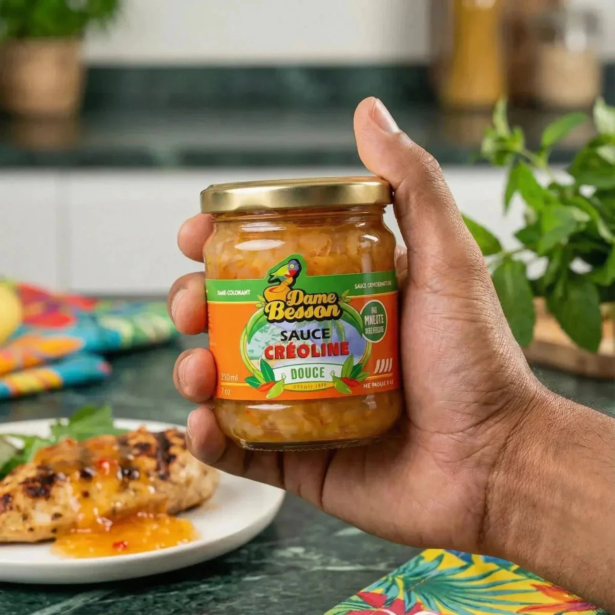 Una mano che tiene un barattolo di salsa creola dolce Dame Besson davanti a un piatto di pollo alla griglia.