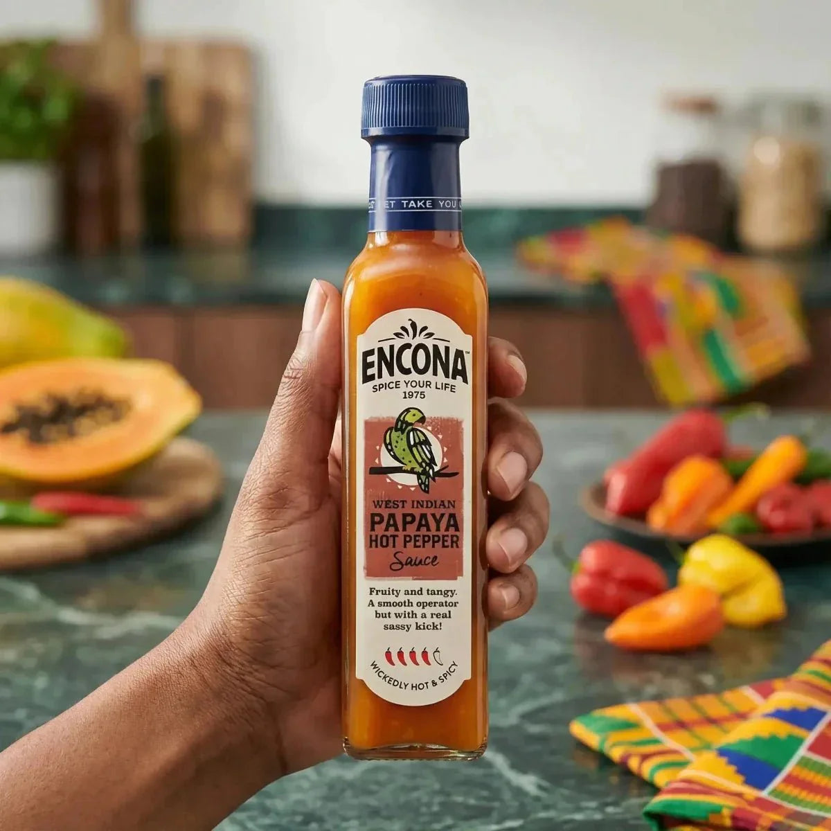 Sauce piquante à la papaye des Antilles par Encona, tenue dans une main devant des papayes.