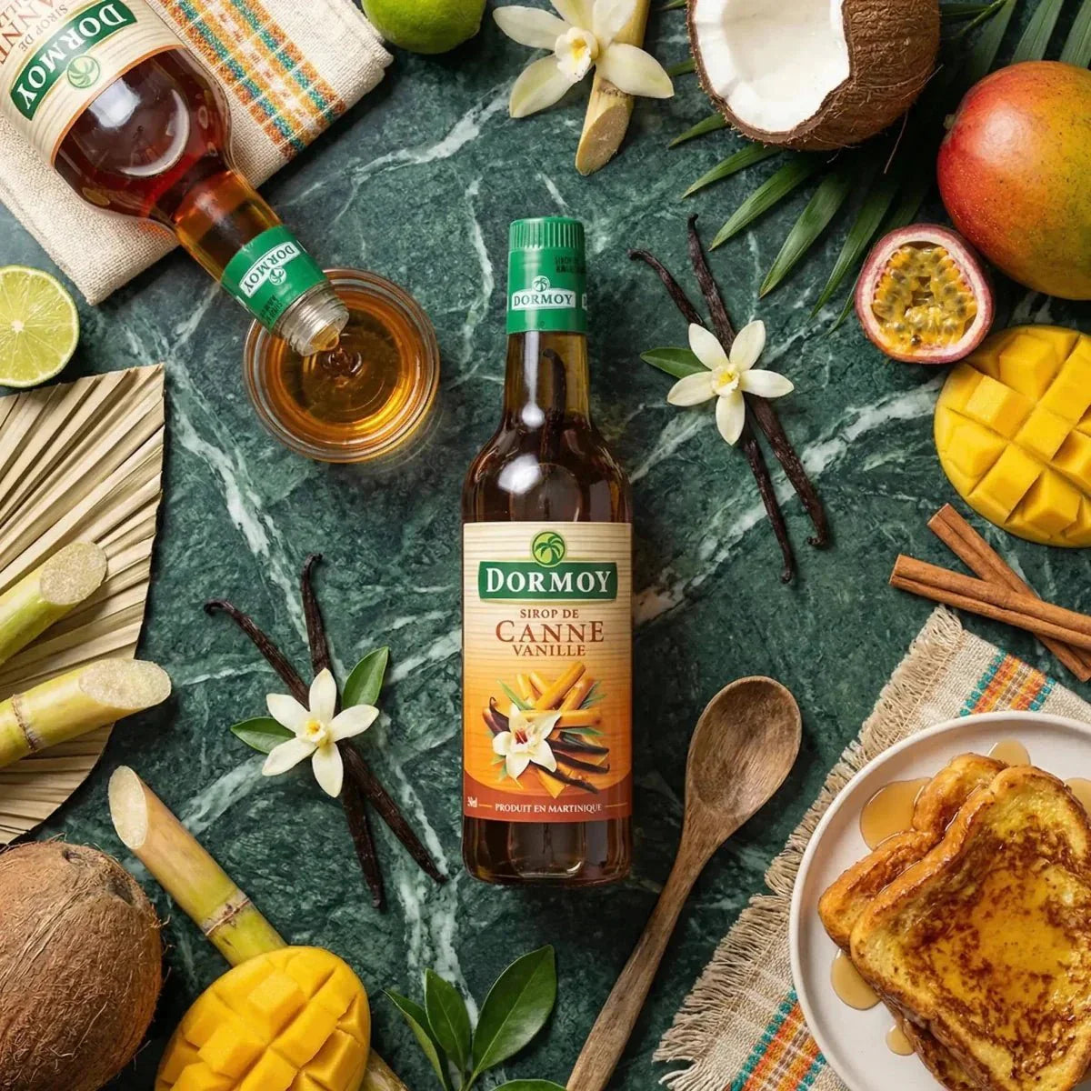 Sciroppo di canna alla vaniglia Dormoy su un tavolo con frutti tropicali, baguette e sciroppo che cola.