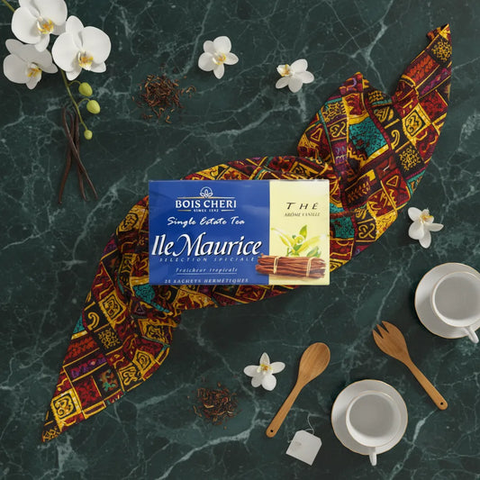 Boîte de thé vanille Bois Chéri posée sur un tissu coloré, entourée d’orchidées, ustensiles et tasses pour une ambiance exotique.